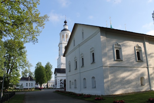 A peaceful setting with a white church featuring traditional architectural elements. The building displays arched windows and a tall bell tower. Lush green trees and a clear blue sky add to the serene atmosphere. There is a pathway and manicured garden beds in the foreground.