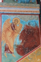A religious mural depicting an angel with large, detailed wings and a halo, holding an open book. The angel is facing a dark, mythical creature with red feathers and horns, seated below. The background features ornate patterns and script in warm colors.