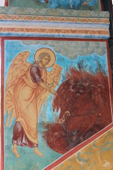 A religious mural depicting an angel with large, detailed wings and a halo, holding an open book. The angel is facing a dark, mythical creature with red feathers and horns, seated below. The background features ornate patterns and script in warm colors.
