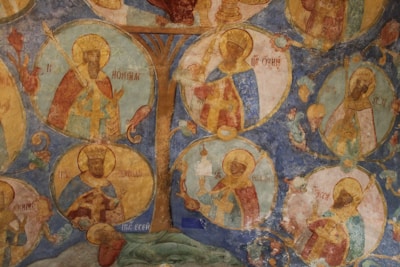 A fresco painting featuring religious figures within circular frames, arranged around a central tree motif. The figures are depicted in traditional attire, with halos and holding various objects. Faded colors and texture indicate an aged artwork.