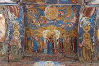 A richly painted religious fresco covers a curved ceiling. The central scene features a depiction of a holy figure surrounded by a group of figures with halos, possibly representing saints or apostles. Above them, celestial beings, angels, and other figures ascend towards a round, golden depiction of a holy figure in a seated position, surrounded by an aura of light with more heavenly figures. The fresco is adorned with intricate details, colorful garments, and vivid expressions, set against a deep blue sky with golden and red accents.