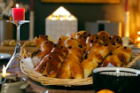 A woven basket is filled with baked goods shaped like small figures, each adorned with raisins for eyes. The table is set for a cozy meal with a lit red candle on a tall candle holder and a warm ambiance created by the glow of additional candles in the background.