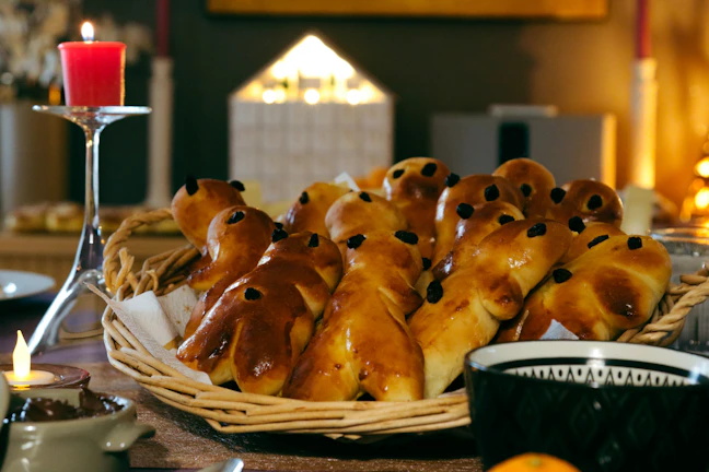 A woven basket is filled with baked goods shaped like small figures, each adorned with raisins for eyes. The table is set for a cozy meal with a lit red candle on a tall candle holder and a warm ambiance created by the glow of additional candles in the background.