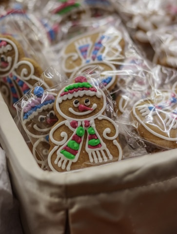 Gingerbread cookies decorated with colorful icing, featuring smiling faces and patterns like scarves and hats. Each cookie is individually wrapped in clear plastic, likely set in a basket.