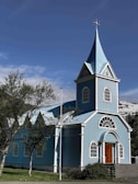 A picturesque blue church with white accents and a steep steeple located in a scenic setting. The church features arched windows and a prominent wooden door. Surrounding the building are green trees and a flagpole, with snow-capped mountains in the background under a clear sky.