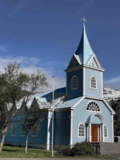 A picturesque blue church with white accents and a steep steeple located in a scenic setting. The church features arched windows and a prominent wooden door. Surrounding the building are green trees and a flagpole, with snow-capped mountains in the background under a clear sky.