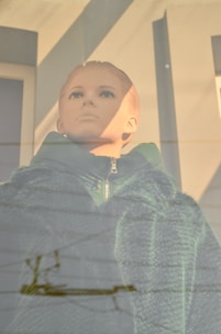 A mannequin with a serene expression is dressed in a green zip-up jacket and a faux fur overcoat. The figure is situated in a display window with light and reflections creating patterns on the surface.