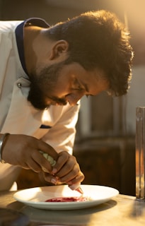 A chef is intently focused on garnishing a dish with a red sauce on a white plate. The scene is warmly lit, creating a professional kitchen ambiance.