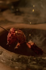A skewer with marinated, grill-cooked meat, possibly tandoori, is suspended over a cooking area. Smoky wisps rise from the meat, which glistens with oil and spices under warm lighting.
