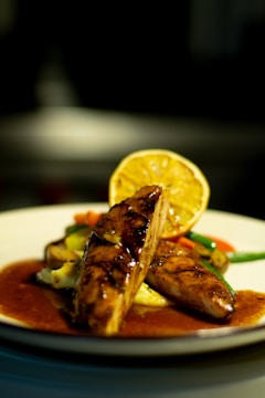 A gourmet dish featuring grilled or roasted meat garnished with a slice of lemon, presented on a white plate. The meat is glazed with a shiny sauce, accompanied by a serving of colorful vegetables including carrots and green beans.