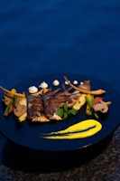 A beautifully plated dish featuring grilled fish accompanied by a variety of colorful vegetables such as mushrooms, carrots, and green beans. A vibrant yellow sauce is artistically smeared on a dark blue plate, adding a touch of elegance and contrast.