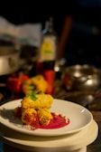 A plated gourmet dish featuring golden-brown pieces garnished with herbs, placed on a vibrant pink sauce with decorative elements on a white plate. The background includes assorted kitchen items slightly out of focus.