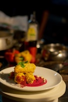 A plated gourmet dish featuring golden-brown pieces garnished with herbs, placed on a vibrant pink sauce with decorative elements on a white plate. The background includes assorted kitchen items slightly out of focus.
