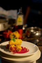 A plated gourmet dish featuring golden-brown pieces garnished with herbs, placed on a vibrant pink sauce with decorative elements on a white plate. The background includes assorted kitchen items slightly out of focus.