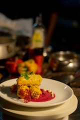 A plated gourmet dish featuring golden-brown pieces garnished with herbs, placed on a vibrant pink sauce with decorative elements on a white plate. The background includes assorted kitchen items slightly out of focus.