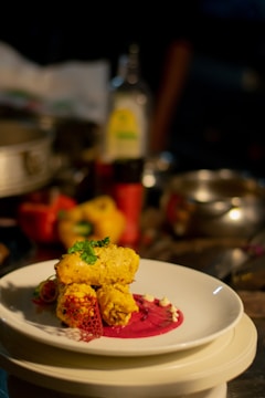 A plated gourmet dish featuring golden-brown pieces garnished with herbs, placed on a vibrant pink sauce with decorative elements on a white plate. The background includes assorted kitchen items slightly out of focus.