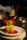 A plated gourmet dish featuring golden-brown pieces garnished with herbs, placed on a vibrant pink sauce with decorative elements on a white plate. The background includes assorted kitchen items slightly out of focus.