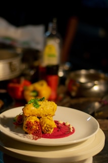 A plated gourmet dish featuring golden-brown pieces garnished with herbs, placed on a vibrant pink sauce with decorative elements on a white plate. The background includes assorted kitchen items slightly out of focus.
