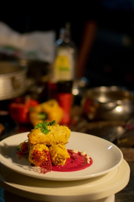 A plated gourmet dish featuring golden-brown pieces garnished with herbs, placed on a vibrant pink sauce with decorative elements on a white plate. The background includes assorted kitchen items slightly out of focus.