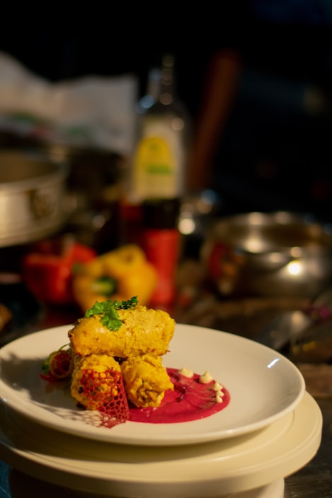 A plated gourmet dish featuring golden-brown pieces garnished with herbs, placed on a vibrant pink sauce with decorative elements on a white plate. The background includes assorted kitchen items slightly out of focus.