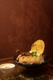 A round dish placed on a black plate contains a baked bread bowl filled with colorful rice, garnished with a meat piece, vegetables, and herbs. A crispy flatbread and garnishes, including lemon wedges, are placed on top. A small cup of yogurt with spices is placed next to the plate. The background is a reddish-brown textured wall.