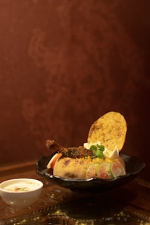 A round dish placed on a black plate contains a baked bread bowl filled with colorful rice, garnished with a meat piece, vegetables, and herbs. A crispy flatbread and garnishes, including lemon wedges, are placed on top. A small cup of yogurt with spices is placed next to the plate. The background is a reddish-brown textured wall.