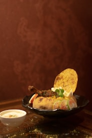 A round dish placed on a black plate contains a baked bread bowl filled with colorful rice, garnished with a meat piece, vegetables, and herbs. A crispy flatbread and garnishes, including lemon wedges, are placed on top. A small cup of yogurt with spices is placed next to the plate. The background is a reddish-brown textured wall.