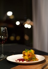 A gourmet dish presented on a white plate, featuring a colorful arrangement of food, including what appears to be seasoned pieces garnished with greens and thinly sliced vegetables. The plate is complemented by artistic streaks of sauce, and in the background, there's a glass of red wine. The setting suggests an elegant dining ambiance with soft lighting and blurred, warm bokeh from lights.