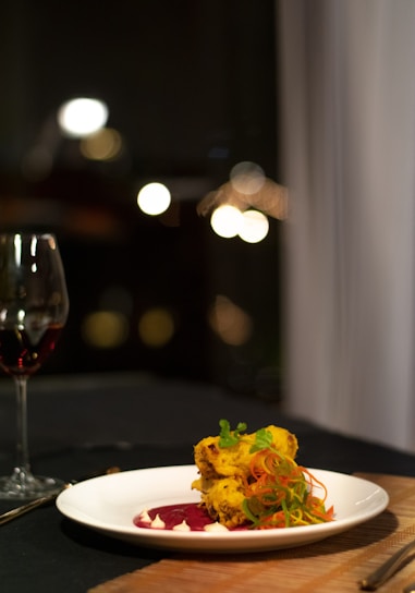A gourmet dish presented on a white plate, featuring a colorful arrangement of food, including what appears to be seasoned pieces garnished with greens and thinly sliced vegetables. The plate is complemented by artistic streaks of sauce, and in the background, there's a glass of red wine. The setting suggests an elegant dining ambiance with soft lighting and blurred, warm bokeh from lights.