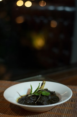 A white bowl filled with a dark, richly textured dish, garnished with fresh green herbs and a stick-like garnish. The bowl sits on a bamboo mat, and the background is softly blurred with warm, bokeh lights.