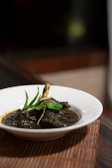 A white bowl containing a rich, dark-colored curry with a bone and garnished with fresh green herbs, possibly mint, set on a woven placemat. The background is softly blurred, highlighting the dish.