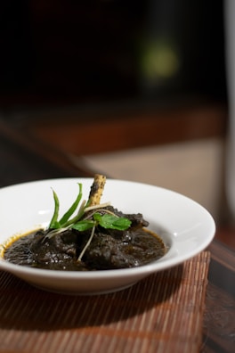 A white bowl containing a rich, dark-colored curry with a bone and garnished with fresh green herbs, possibly mint, set on a woven placemat. The background is softly blurred, highlighting the dish.