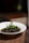 A white bowl containing a rich, dark-colored curry with a bone and garnished with fresh green herbs, possibly mint, set on a woven placemat. The background is softly blurred, highlighting the dish.