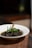 A white bowl containing a rich, dark-colored curry with a bone and garnished with fresh green herbs, possibly mint, set on a woven placemat. The background is softly blurred, highlighting the dish.