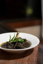 A white bowl containing a rich, dark-colored curry with a bone and garnished with fresh green herbs, possibly mint, set on a woven placemat. The background is softly blurred, highlighting the dish.