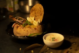 A vibrant dish featuring biryani served in a round bread bowl. A piece of cooked chicken with onion rings and garnishes is placed on top. A papadum is balanced upright, adding a crispy element. A small bowl of yogurt with herbs and spices is beside it, all set on a dark, textured surface.
