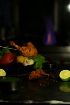 A piece of grilled meat, possibly chicken, is positioned prominently on a skewer with a backdrop of fresh vegetables including a tomato, lime, and green garnish. A small container with green sauce is visible on the metallic surface. A blue flame is faintly visible in the background, suggesting it might be in a kitchen or restaurant setting.