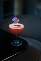A cocktail in a coupe glass sits on a dark reflective surface. The drink is red with a foamy top, garnished with a purple flower and a small cinnamon stick. The background appears softly blurred, contributing to a focus on the drink.