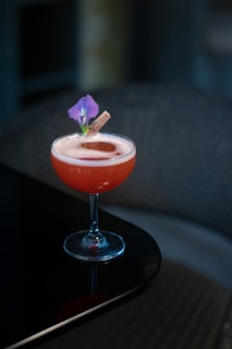 A cocktail in a coupe glass sits on a dark reflective surface. The drink is red with a foamy top, garnished with a purple flower and a small cinnamon stick. The background appears softly blurred, contributing to a focus on the drink.