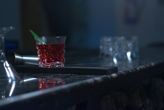 A dimly lit bar counter featuring a glass filled with a red beverage, a green stirrer, and a metal shaker beside it. In the background, there are several blurred glasses, contributing to a moody atmosphere.