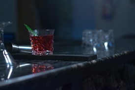 A dimly lit bar counter featuring a glass filled with a red beverage, a green stirrer, and a metal shaker beside it. In the background, there are several blurred glasses, contributing to a moody atmosphere.