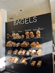 A black wall with the word 'BAGELS' at the top in large white letters. Below, multiple wire baskets are mounted on the wall, each filled with a variety of bagels. The bagels vary in types, such as poppy, sesame, rye, wheat, multigrain, spinach, asiago, jalapeño, and everything. Natural light illuminates the upper left of the display, creating a warm and inviting atmosphere.