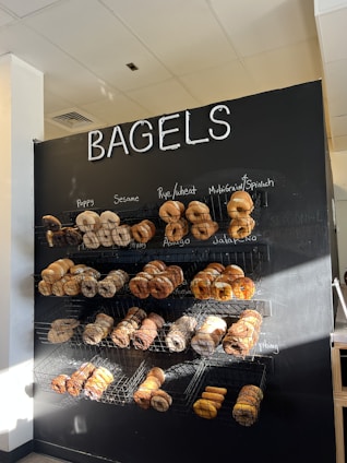 A black wall with the word 'BAGELS' at the top in large white letters. Below, multiple wire baskets are mounted on the wall, each filled with a variety of bagels. The bagels vary in types, such as poppy, sesame, rye, wheat, multigrain, spinach, asiago, jalapeño, and everything. Natural light illuminates the upper left of the display, creating a warm and inviting atmosphere.