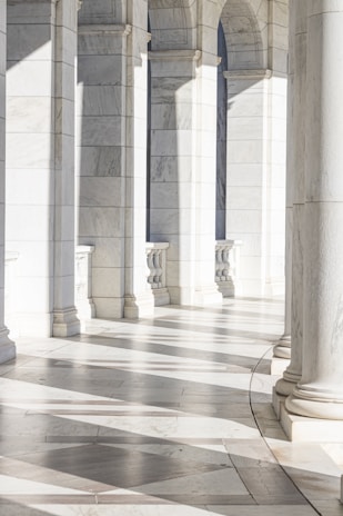 Elegant marble arches cast intricate shadows on the smooth tiled floor, creating a serene and sophisticated pattern. The space appears bright and open, with natural light enhancing the architectural details.