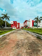 A modern building with a bold red and white facade is surrounded by palm trees and manicured lawns. The sky is lightly clouded, and the wide pathway leading to the building suggests an institutional or corporate setting.