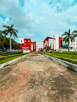 A modern building with a bold red and white facade is surrounded by palm trees and manicured lawns. The sky is lightly clouded, and the wide pathway leading to the building suggests an institutional or corporate setting.