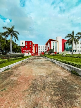 A modern building with a bold red and white facade is surrounded by palm trees and manicured lawns. The sky is lightly clouded, and the wide pathway leading to the building suggests an institutional or corporate setting.