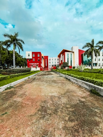 A modern building with a bold red and white facade is surrounded by palm trees and manicured lawns. The sky is lightly clouded, and the wide pathway leading to the building suggests an institutional or corporate setting.