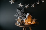 A cozy, rustic holiday scene with small decorative house shapes and star-shaped lights as the centerpiece. The houses have lit interiors, glowing warmly. A large star and snowflake decorations frame the scene on a dark background.