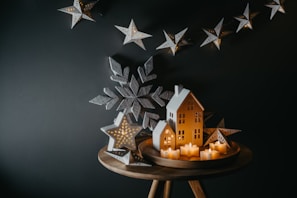 A cozy, rustic holiday scene with small decorative house shapes and star-shaped lights as the centerpiece. The houses have lit interiors, glowing warmly. A large star and snowflake decorations frame the scene on a dark background.